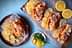 Four lobster rolls served with potato chips and lemon slices on a wooden platter. - Olive Oil Times
