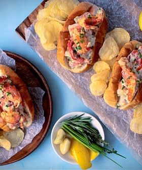 Four lobster rolls served with potato chips and lemon slices on a wooden platter. - Olive Oil Times