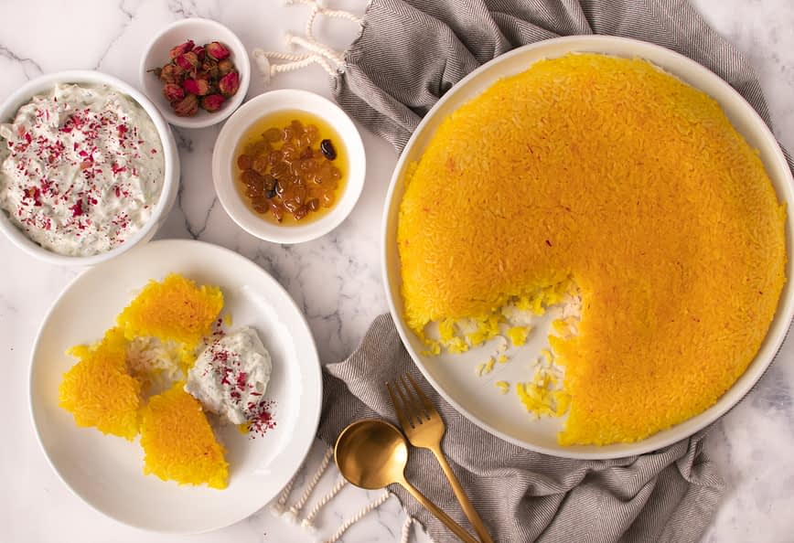 A round saffron rice dessert with a portion removed, served with yogurt and dried fruits. - Olive Oil Times