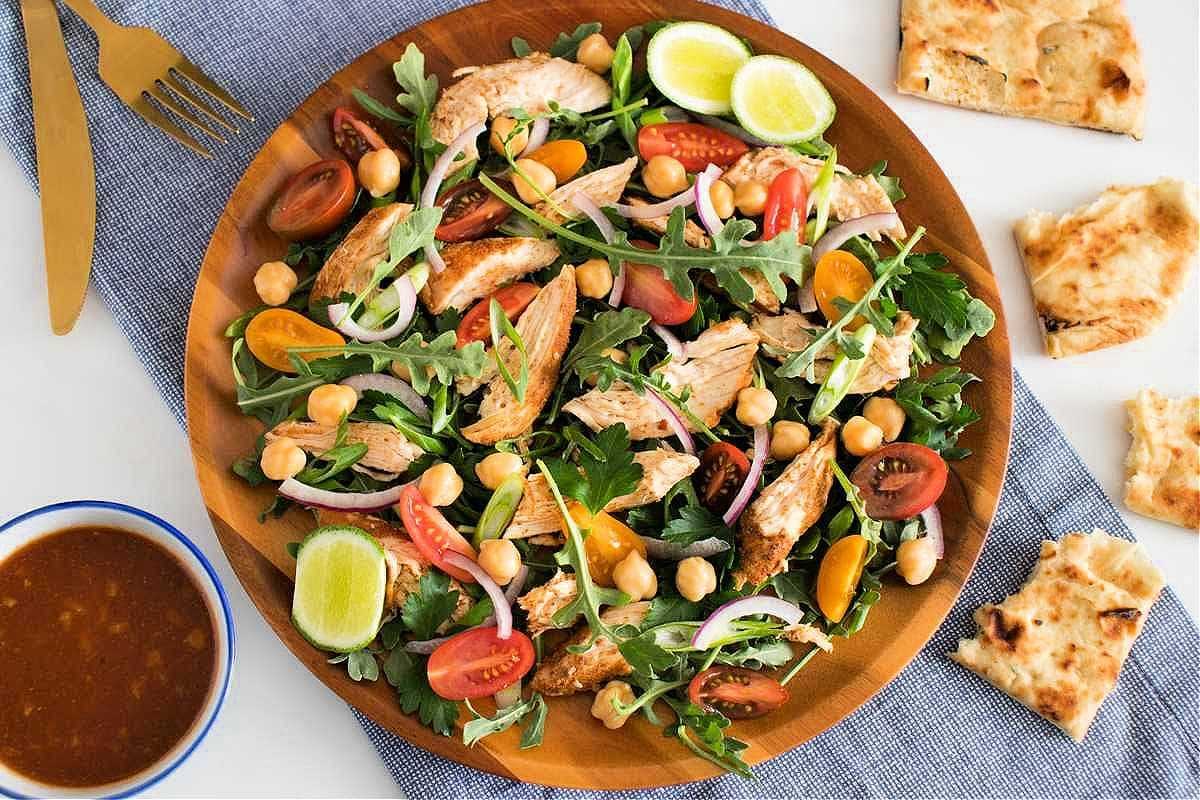 A large wooden bowl filled with a mixed salad featuring grilled chicken, arugula, tomatoes, and chickpeas. - Olive Oil Times