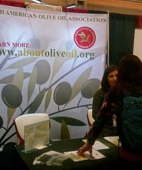 A booth for the North American Olive Oil Association with a banner and a person interacting with staff. - Olive Oil Times