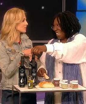 Two women interacting while presenting various olive oil products on a table. - Olive Oil Times