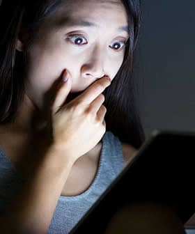 A woman with long hair looking shocked while holding a tablet in a dimly lit room. - Olive Oil Times