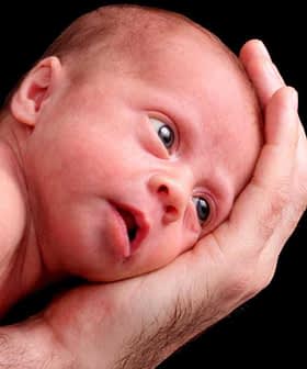 A newborn baby resting on an adult's hand with a neutral expression and wide-open eyes. - Olive Oil Times