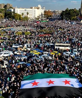 A large crowd gathered in a public square holding a Syrian flag with three stars. - Olive Oil Times
