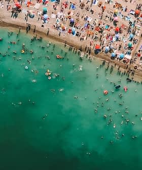 Aerial view of a crowded beach with swimmers and colorful umbrellas along the shoreline. - Olive Oil Times